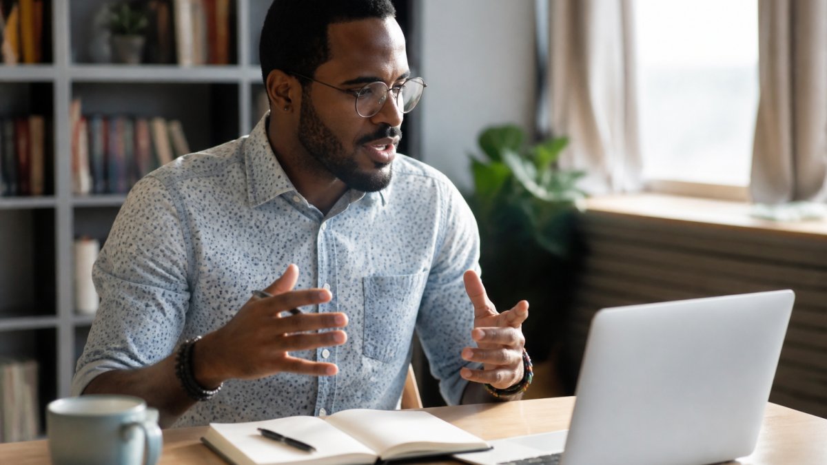 A business professional speaking on a video call, gesturing toward a laptop screen showing a presentation, with a notebook and coffee on a clean desk in a home office